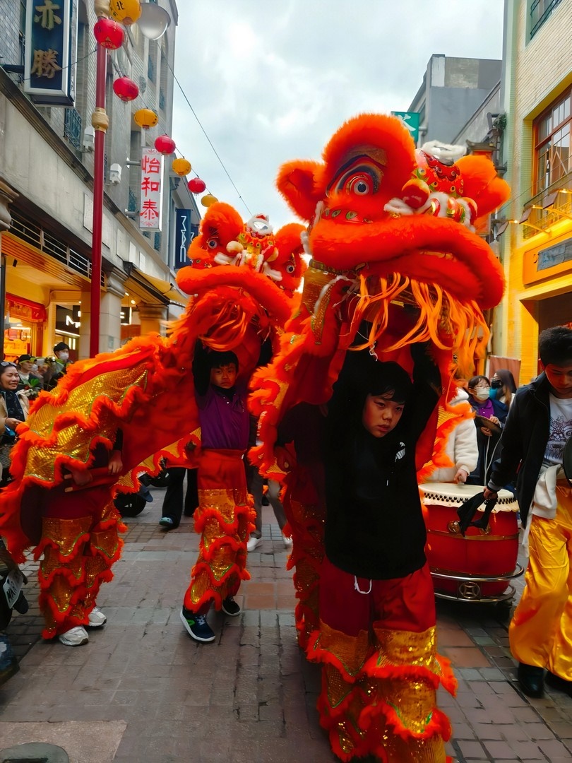 學生們神情專注地操控色彩鮮豔、栩栩如生的紅色獅頭與獅尾，在在富有歷史氛圍的迪化商圈熱鬧街道間舞動穿梭,展現出充滿活力的精湛技巧。不僅讓學生們有機會將所學展現在社區,也促進了傳統文化的傳承與在地連結。
